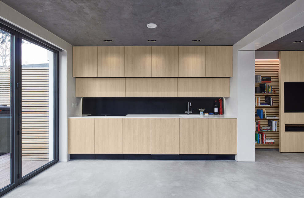 This is an example of a small contemporary single-wall kitchen in London with an undermount sink, flat-panel cabinets, black splashback, panelled appliances, concrete floors, no island, grey floor, light wood cabinets and white benchtop.