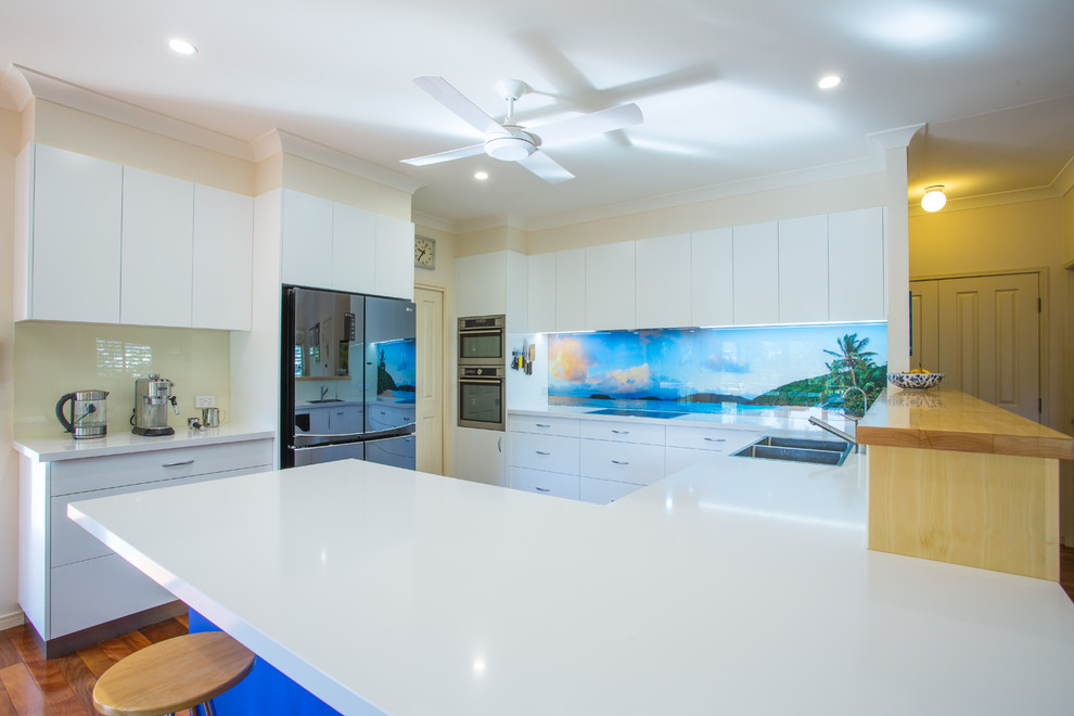 Design ideas for a mid-sized beach style u-shaped kitchen pantry in Brisbane with a double-bowl sink, flat-panel cabinets, blue cabinets, quartz benchtops, blue splashback, glass sheet splashback, stainless steel appliances, dark hardwood floors, with island, brown floor and white benchtop.