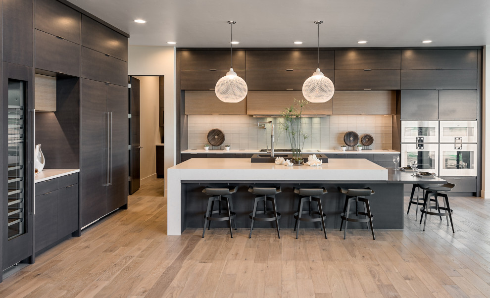 Example of a trendy l-shaped light wood floor and beige floor kitchen design in Miami with flat-panel cabinets, dark wood cabinets, gray backsplash, stainless steel appliances, an island and white countertops