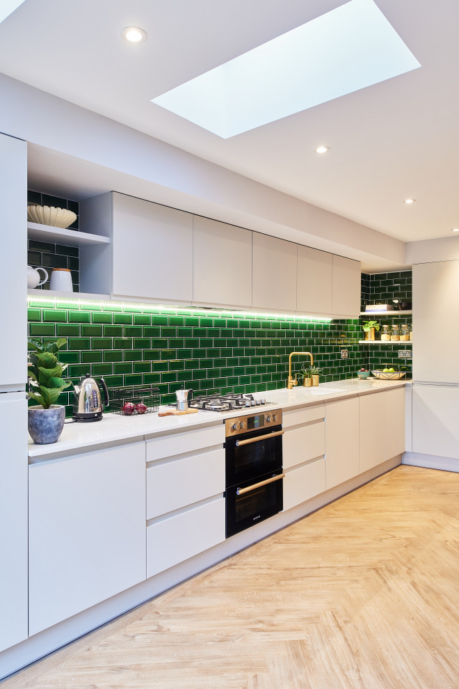 This is an example of a mid-sized contemporary l-shaped open plan kitchen with an integrated sink, flat-panel cabinets, white cabinets, solid surface benchtops, green splashback, subway tile splashback, panelled appliances, vinyl floors, no island, multi-coloured floor and white benchtop.