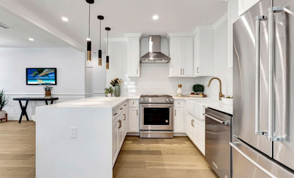 Contemporary u-shaped kitchen in Los Angeles with a belfast sink, shaker cabinets, white cabinets, composite countertops, white splashback, metro tiled splashback, stainless steel appliances, medium hardwood flooring, an island, brown floors and white worktops.