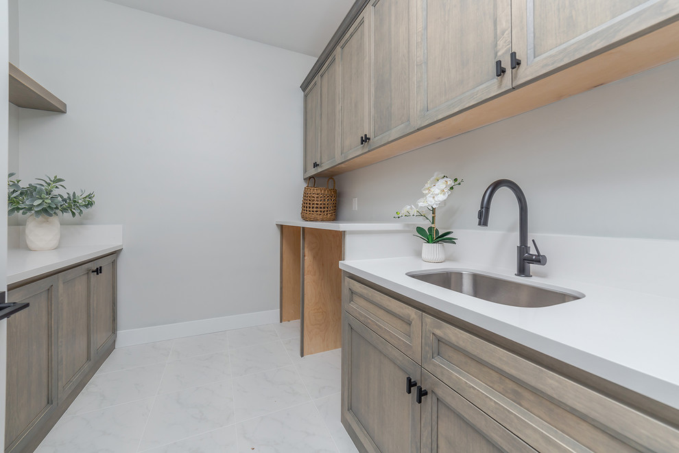This is an example of a large country u-shaped dedicated laundry room in Sacramento with an integrated sink, flat-panel cabinets, medium wood cabinets, solid surface benchtops, grey walls, ceramic floors, an integrated washer and dryer, white floor and white benchtop.