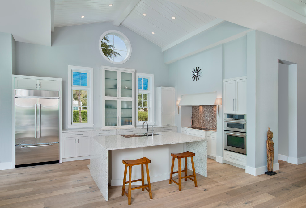 This is an example of a beach style l-shaped kitchen in Tampa with glass-front cabinets, white cabinets, stainless steel appliances, with island, an undermount sink, multi-coloured splashback, mosaic tile splashback and light hardwood floors.