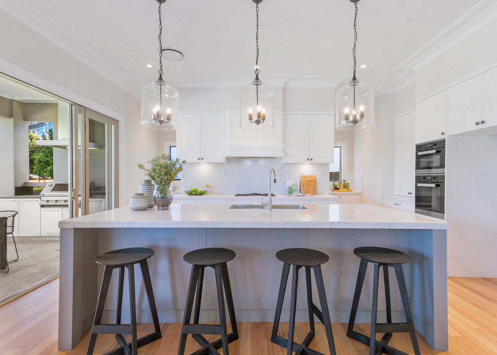 Inspiration for a mid-sized transitional galley open plan kitchen in Sydney with a double-bowl sink, shaker cabinets, white cabinets, marble benchtops, marble splashback, stainless steel appliances, light hardwood floors, with island and white benchtop.