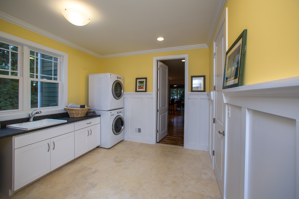 Photo of a large arts and crafts single-wall utility room in DC Metro with shaker cabinets, white cabinets, granite benchtops, yellow walls, porcelain floors, a stacked washer and dryer and a drop-in sink.