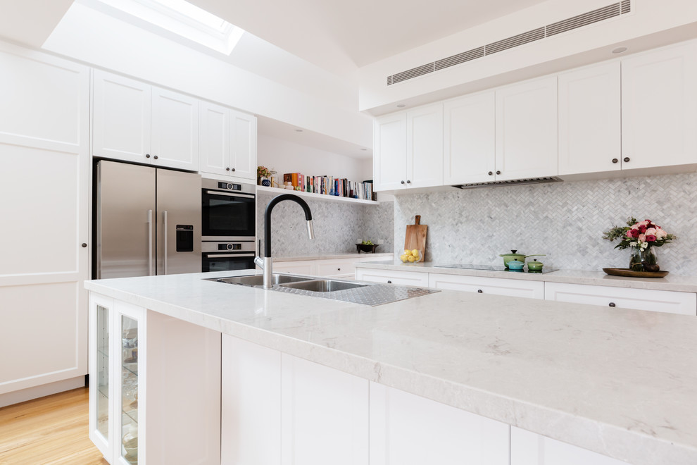Large transitional l-shaped open plan kitchen in Melbourne with a double-bowl sink, shaker cabinets, white cabinets, quartz benchtops, grey splashback, mosaic tile splashback, stainless steel appliances, light hardwood floors, with island and brown floor.