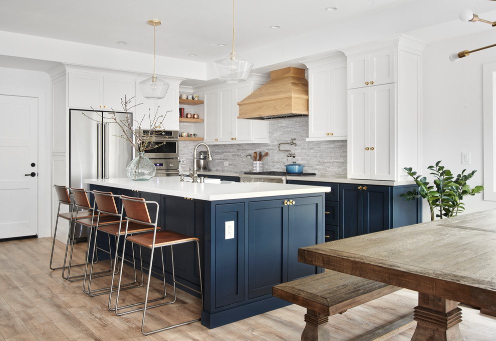 Photo of a large beach style l-shaped open plan kitchen in San Francisco with shaker cabinets, grey splashback, stainless steel appliances, light hardwood floors, with island, a farmhouse sink, white cabinets, solid surface benchtops, brown floor and white benchtop.