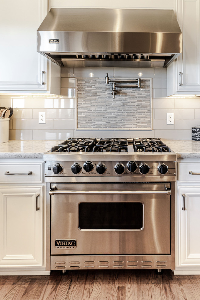 Exempel på ett stort lantligt vit vitt kök, med en undermonterad diskho, luckor med upphöjd panel, vita skåp, bänkskiva i kvarts, grått stänkskydd, stänkskydd i glaskakel, rostfria vitvaror, ljust trägolv, en halv köksö och brunt golv