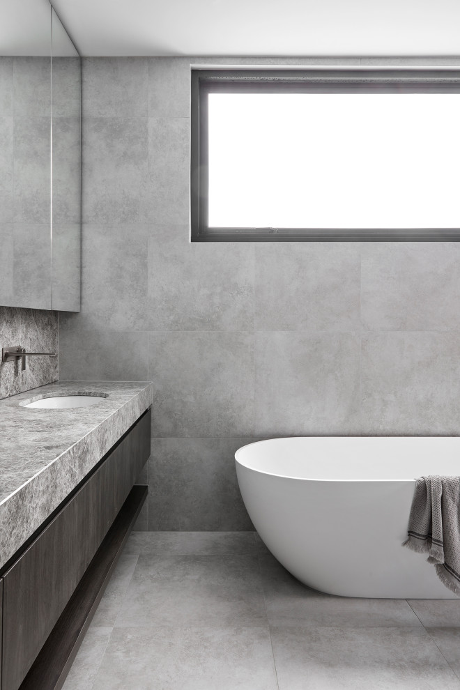 Пример оригинального дизайна: главная ванная комната среднего размера в стиле модернизм с фасадами островного типа, темными деревянными фасадами, отдельно стоящей ванной, душем в нише, унитазом-моноблоком, серой плиткой, керамической плиткой, серыми стенами, полом из керамической плитки, монолитной раковиной, столешницей из известняка, серым полом, душем с распашными дверями, серой столешницей, нишей, тумбой под две раковины и встроенной тумбой
