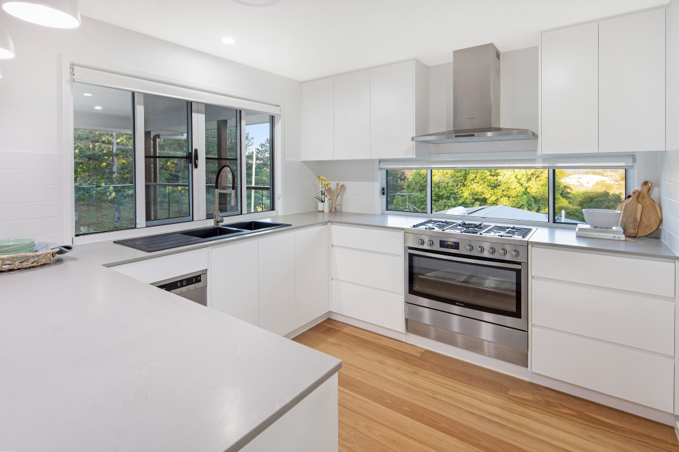 Photo of a mid-sized contemporary u-shaped kitchen in Sunshine Coast with a double-bowl sink, white cabinets, concrete benchtops, white splashback, subway tile splashback, stainless steel appliances, no island, flat-panel cabinets, medium hardwood floors, brown floor and grey benchtop.