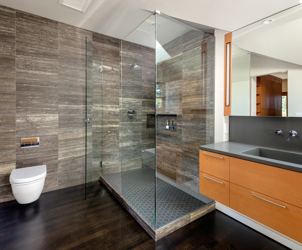 Photo of a mid-sized modern master bathroom in Portland with flat-panel cabinets, medium wood cabinets, a curbless shower, a wall-mount toilet, gray tile, travertine, white walls, dark hardwood floors, an integrated sink, engineered quartz benchtops, black floor and a hinged shower door.