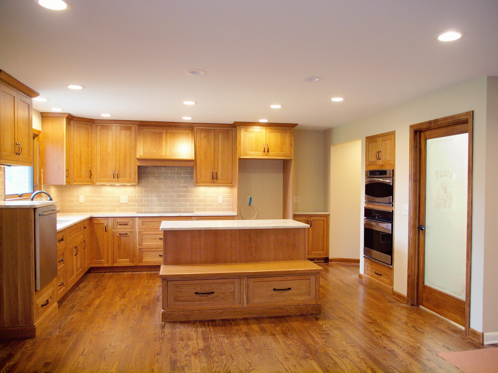 This is an example of a mid-sized traditional l-shaped eat-in kitchen in Cincinnati with an undermount sink, shaker cabinets, medium wood cabinets, solid surface benchtops, beige splashback, porcelain splashback, stainless steel appliances, dark hardwood floors, with island and brown floor.