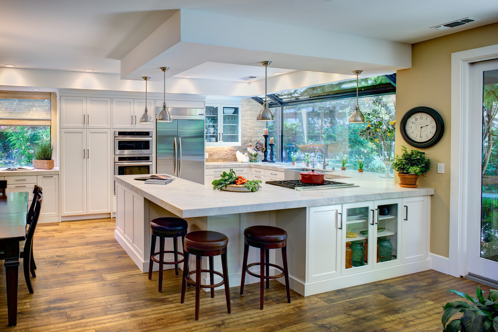 Design ideas for a mid-sized transitional u-shaped open plan kitchen in Sacramento with a farmhouse sink, shaker cabinets, white cabinets, granite benchtops, white splashback, stainless steel appliances, light hardwood floors and a peninsula.