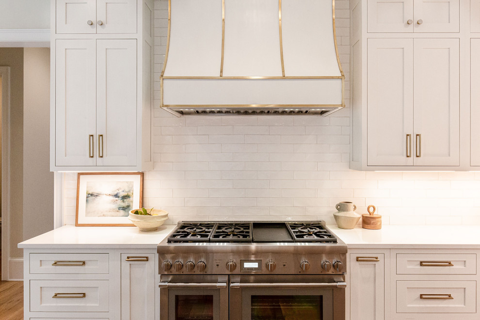 Inredning av ett klassiskt stort vit vitt kök, med en undermonterad diskho, luckor med profilerade fronter, vita skåp, bänkskiva i kvarts, vitt stänkskydd, stänkskydd i tunnelbanekakel, integrerade vitvaror, mellanmörkt trägolv, en köksö och beiget golv