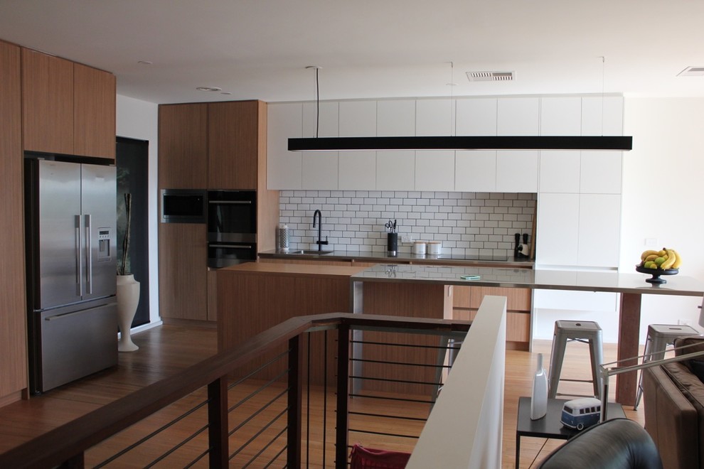 Photo of a mid-sized modern galley open plan kitchen in Melbourne with an undermount sink, flat-panel cabinets, light wood cabinets, stainless steel benchtops, white splashback, subway tile splashback, stainless steel appliances, light hardwood floors and with island.