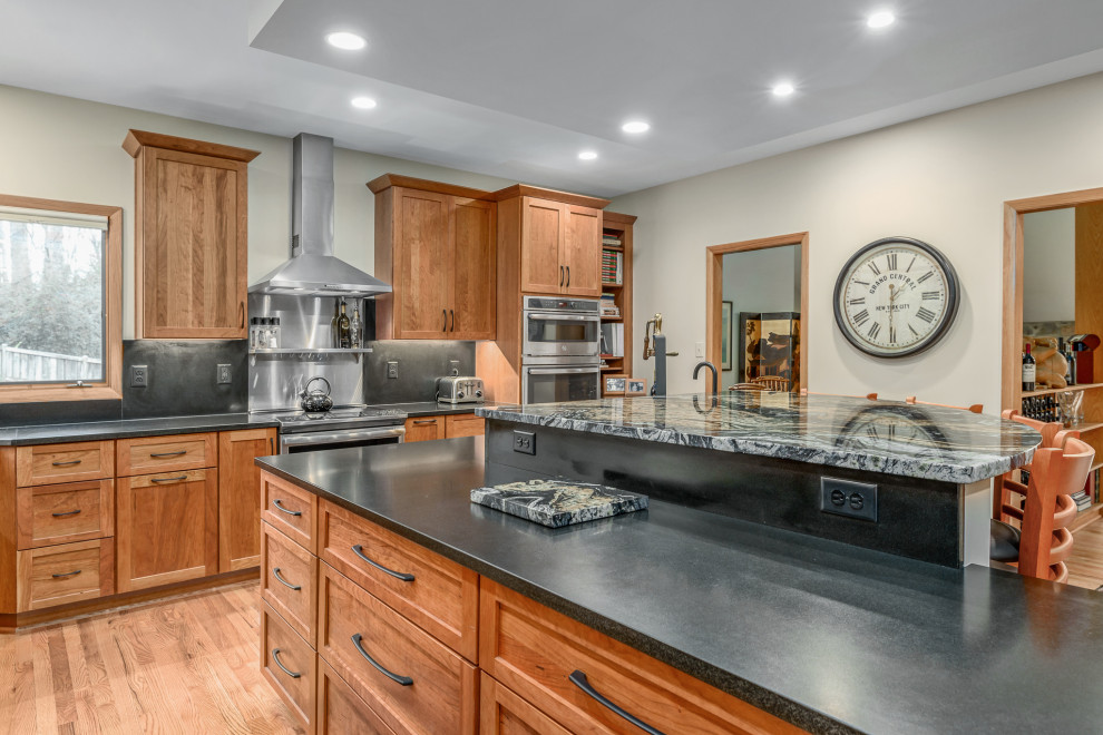 Photo of a transitional l-shaped kitchen in DC Metro with a drop-in sink, recessed-panel cabinets, brown cabinets, granite benchtops, black splashback, ceramic splashback, stainless steel appliances, laminate floors, with island, brown floor and black benchtop.