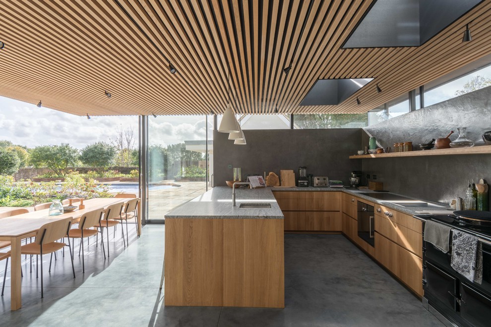 Mid-sized contemporary u-shaped eat-in kitchen in Sussex with an undermount sink, flat-panel cabinets, light wood cabinets, grey splashback, concrete floors, grey floor, a peninsula and black appliances.