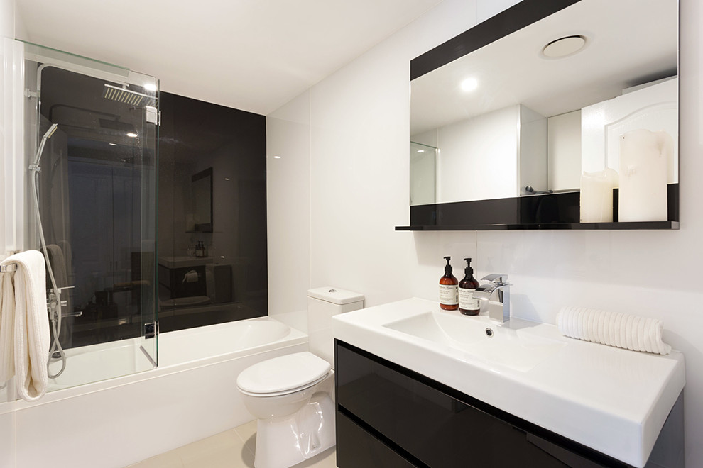 This is an example of a mid-sized contemporary 3/4 bathroom in Brisbane with an alcove tub, a shower/bathtub combo, a wall-mount toilet, beige tile, porcelain tile, white walls, flat-panel cabinets, black cabinets, an integrated sink, beige floor and a hinged shower door.