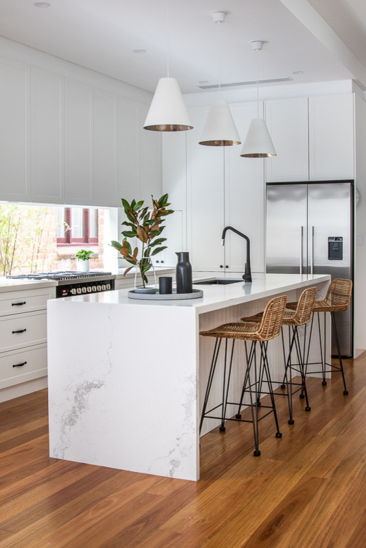 Design ideas for a beach style l-shaped eat-in kitchen in Sydney with an undermount sink, shaker cabinets, white cabinets, window splashback, stainless steel appliances, medium hardwood floors, with island, brown floor and white benchtop.