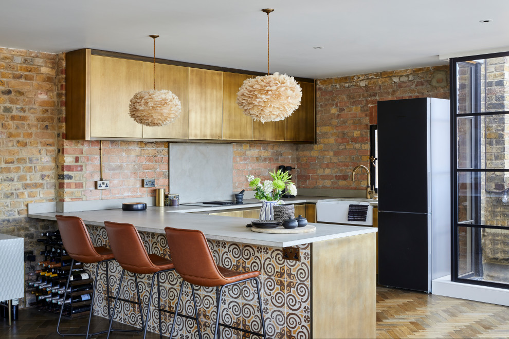 This is an example of a contemporary u-shaped kitchen in London with a farmhouse sink, flat-panel cabinets, red splashback, brick splashback, black appliances, medium hardwood floors, a peninsula, brown floor and white benchtop.