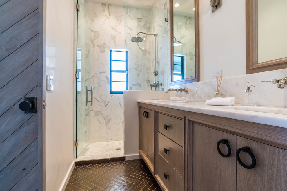 Photo of a small transitional 3/4 bathroom in Miami with recessed-panel cabinets, medium wood cabinets, an alcove shower, white tile, stone slab, white walls, brick floors, an undermount sink, marble benchtops, brown floor, a hinged shower door and white benchtops.