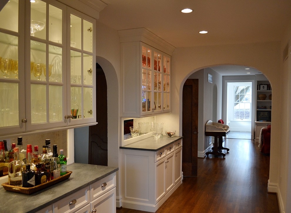 This is an example of a large transitional single-wall wet bar in St Louis with white cabinets, grey splashback, stone tile splashback, medium hardwood floors, glass-front cabinets, no sink and soapstone benchtops.