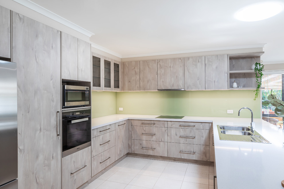 Inspiration for a mid-sized modern u-shaped kitchen pantry in Adelaide with a double-bowl sink, flat-panel cabinets, light wood cabinets, quartz benchtops, green splashback, glass sheet splashback, stainless steel appliances, porcelain floors, a peninsula, white floor and white benchtop.