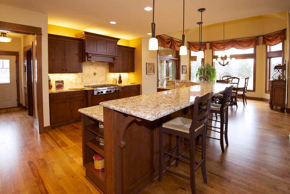 This is an example of a large traditional l-shaped eat-in kitchen in Milwaukee with recessed-panel cabinets, medium wood cabinets, granite benchtops, beige splashback, ceramic splashback, stainless steel appliances, medium hardwood floors, with island, an undermount sink, brown floor and multi-coloured benchtop.