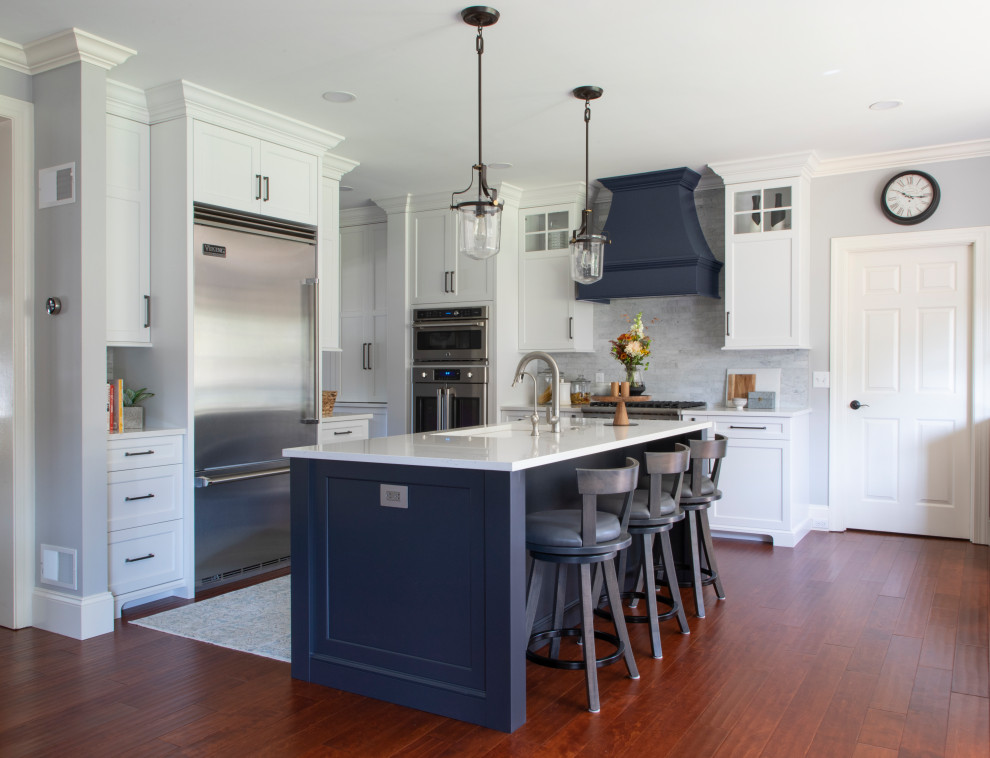 Inspiration for a transitional l-shaped medium tone wood floor and brown floor kitchen remodel with an undermount sink, shaker cabinets, quartz countertops, gray backsplash, porcelain backsplash, stainless steel appliances, an island, white countertops and white cabinets