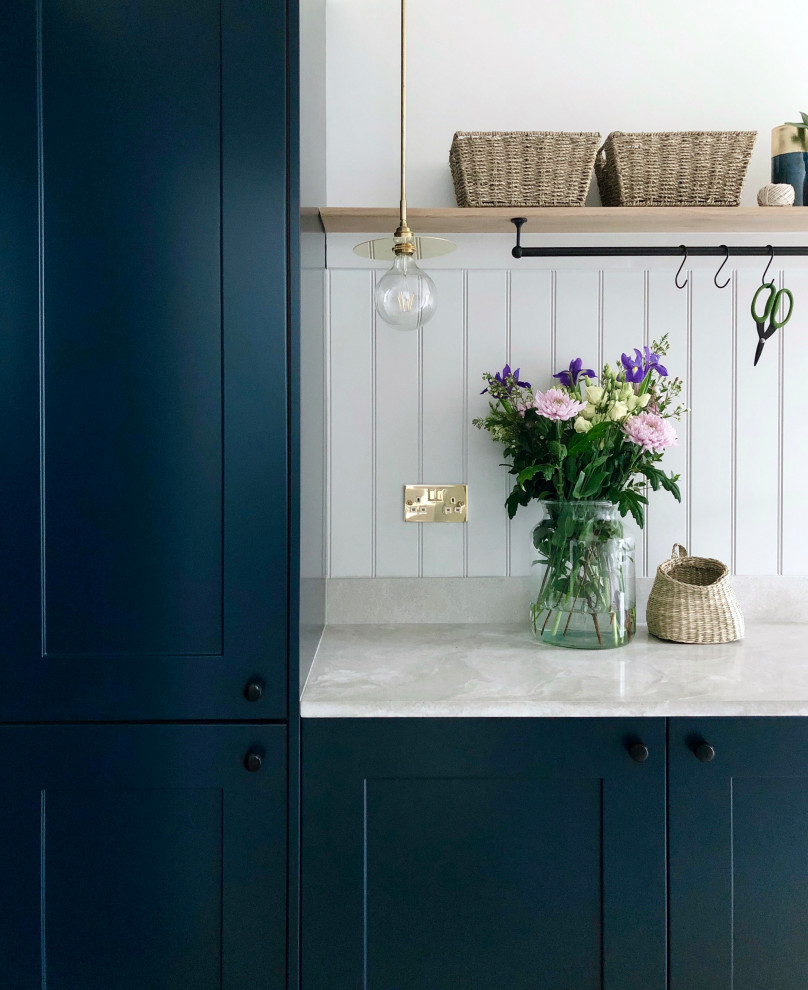 This is an example of a small traditional single-wall utility room with a farmhouse sink, shaker cabinets, blue cabinets, grey walls, laminate floors, an integrated washer and dryer, beige floor, grey benchtop and planked wall panelling.