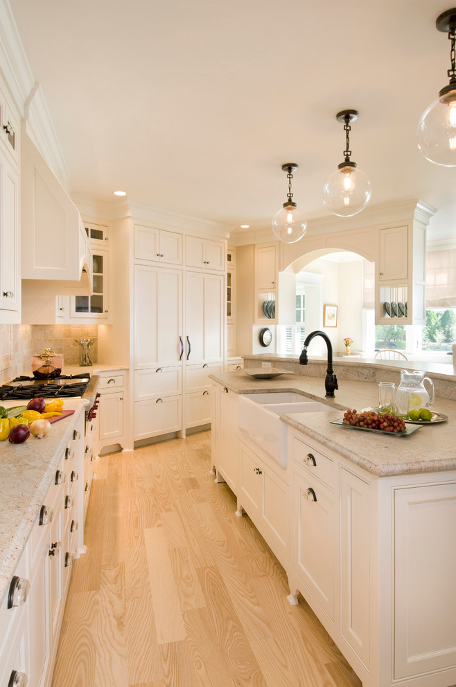 Photo of a large traditional galley kitchen in Boston with a farmhouse sink, white cabinets, beige splashback, panelled appliances, shaker cabinets, granite benchtops, ceramic splashback, light hardwood floors and with island.