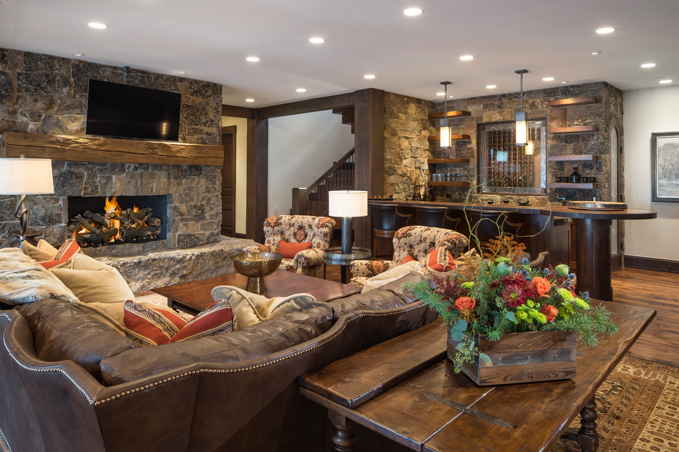 Inspiration for a large country open concept family room in Salt Lake City with white walls, medium hardwood floors, a standard fireplace, a stone fireplace surround, brown floor, a wall-mounted tv and a home bar.