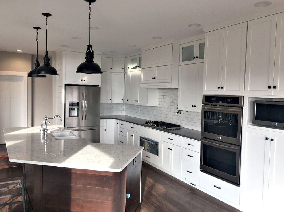 This is an example of a mid-sized transitional l-shaped open plan kitchen in Other with a farmhouse sink, shaker cabinets, white cabinets, quartzite benchtops, white splashback, subway tile splashback, stainless steel appliances, dark hardwood floors, with island and brown floor.