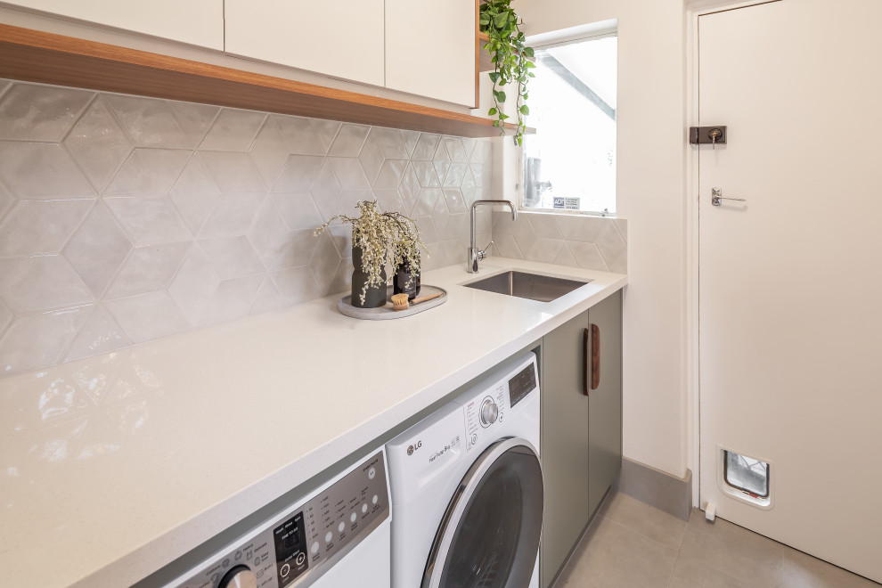 Photo of a small contemporary single-wall separated utility room in Adelaide with a submerged sink, flat-panel cabinets, green cabinets, engineered stone countertops, grey splashback, porcelain splashback, white walls, ceramic flooring, a side by side washer and dryer, grey floors and white worktops.