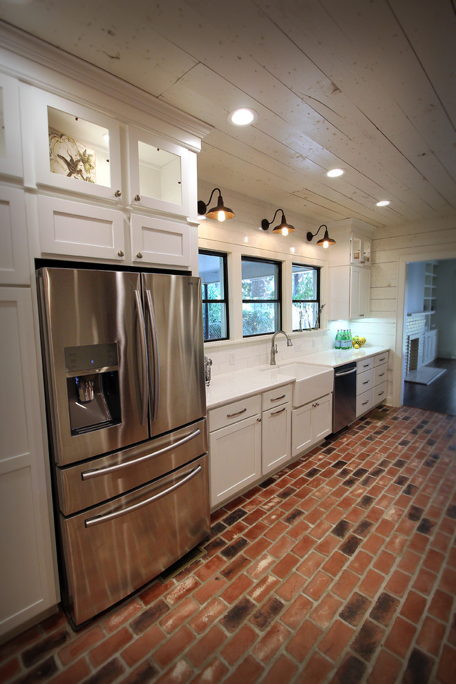 Design ideas for a country l-shaped kitchen in New Orleans with a farmhouse sink, shaker cabinets, white cabinets, quartz benchtops, white splashback, subway tile splashback, stainless steel appliances, brick floors and with island.