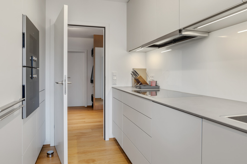Zweizeilige, Kleine Moderne Küche mit Einbauwaschbecken, flächenbündigen Schrankfronten, Küchenrückwand in Weiß, Glasrückwand, schwarzen Elektrogeräten und braunem Holzboden in Nürnberg