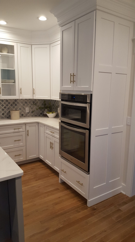 Eat-in kitchen - large transitional u-shaped light wood floor and brown floor eat-in kitchen idea in DC Metro with a farmhouse sink, recessed-panel cabinets, white cabinets, quartzite countertops, gray backsplash, marble backsplash, stainless steel appliances, an island and white countertops