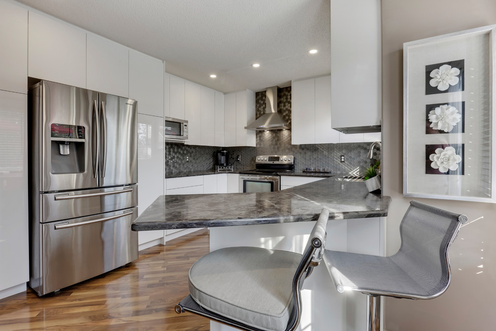 Mid-sized contemporary u-shaped eat-in kitchen in Calgary with an undermount sink, flat-panel cabinets, white cabinets, quartz benchtops, grey splashback, glass tile splashback, stainless steel appliances, laminate floors, a peninsula, brown floor and black benchtop.