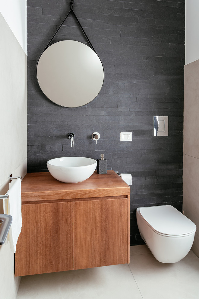 This is an example of a small contemporary powder room in Naples with flat-panel cabinets, medium wood cabinets, a wall-mount toilet, black tile, slate, porcelain floors, a vessel sink, wood benchtops, black walls and grey floor.