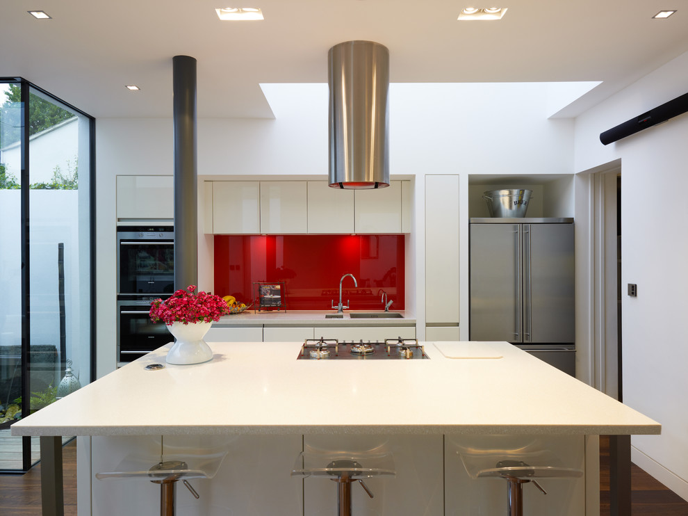 This is an example of a contemporary kitchen in London with an undermount sink, flat-panel cabinets, white cabinets, red splashback, glass sheet splashback, stainless steel appliances and with island.