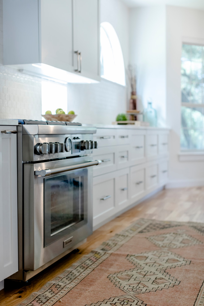 Inspiration for a large transitional l-shaped light wood floor and brown floor eat-in kitchen remodel in Austin with a single-bowl sink, shaker cabinets, white cabinets, quartz countertops, white backsplash, ceramic backsplash, stainless steel appliances, an island and white countertops