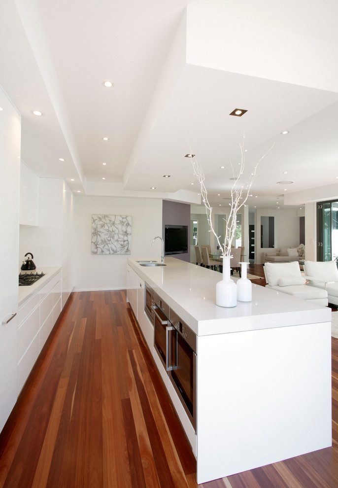Photo of a modern galley eat-in kitchen in Sydney with a drop-in sink, flat-panel cabinets, white cabinets, quartz benchtops, white splashback, glass tile splashback, stainless steel appliances, medium hardwood floors and with island.