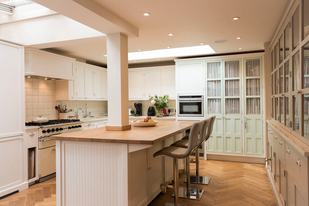 Photo of a large transitional u-shaped eat-in kitchen in London with shaker cabinets, white cabinets, wood benchtops, white splashback, ceramic splashback, medium hardwood floors, with island, brown floor, an undermount sink, stainless steel appliances and beige benchtop.