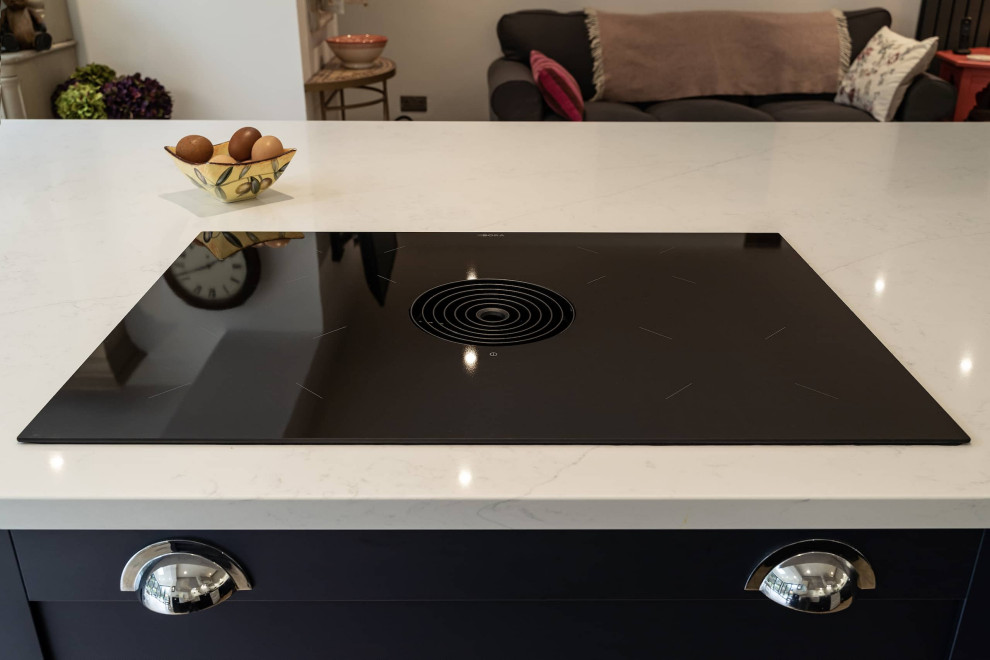 This is an example of a medium sized modern l-shaped kitchen/diner in Oxfordshire with a belfast sink, shaker cabinets, white cabinets, quartz worktops, white splashback, black appliances, an island and white worktops.