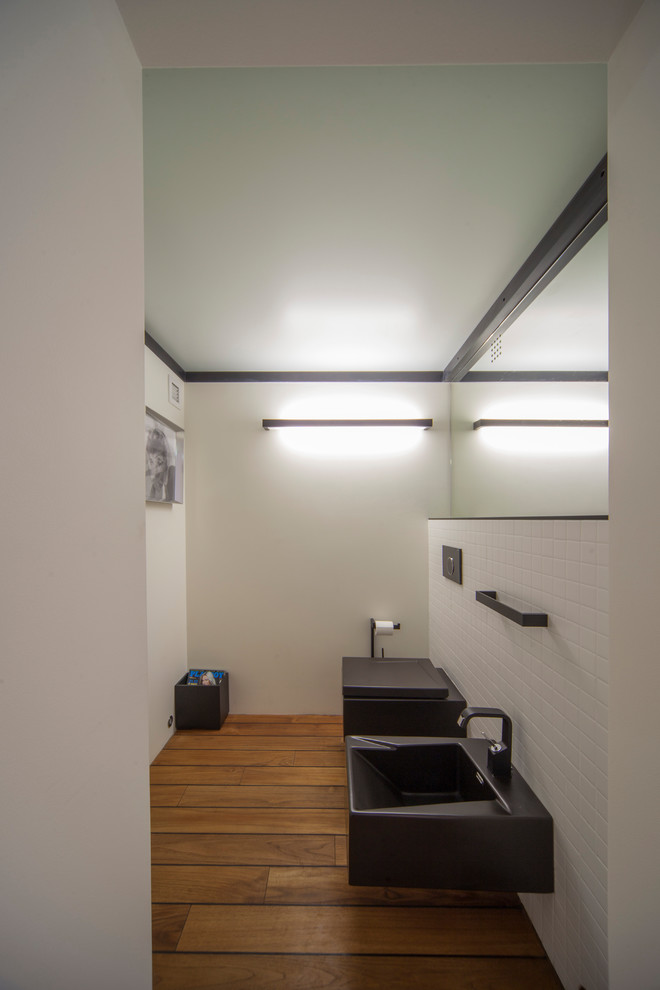 Small contemporary powder room in Moscow with a wall-mount toilet, white tile, ceramic tile, a wall-mount sink, brown floor and medium hardwood floors.