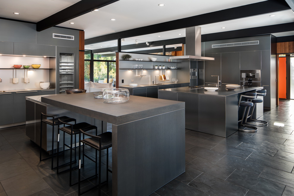 Photo of an expansive contemporary galley kitchen in Los Angeles with flat-panel cabinets, grey cabinets, stainless steel benchtops, white splashback, stainless steel appliances, multiple islands and black floor.