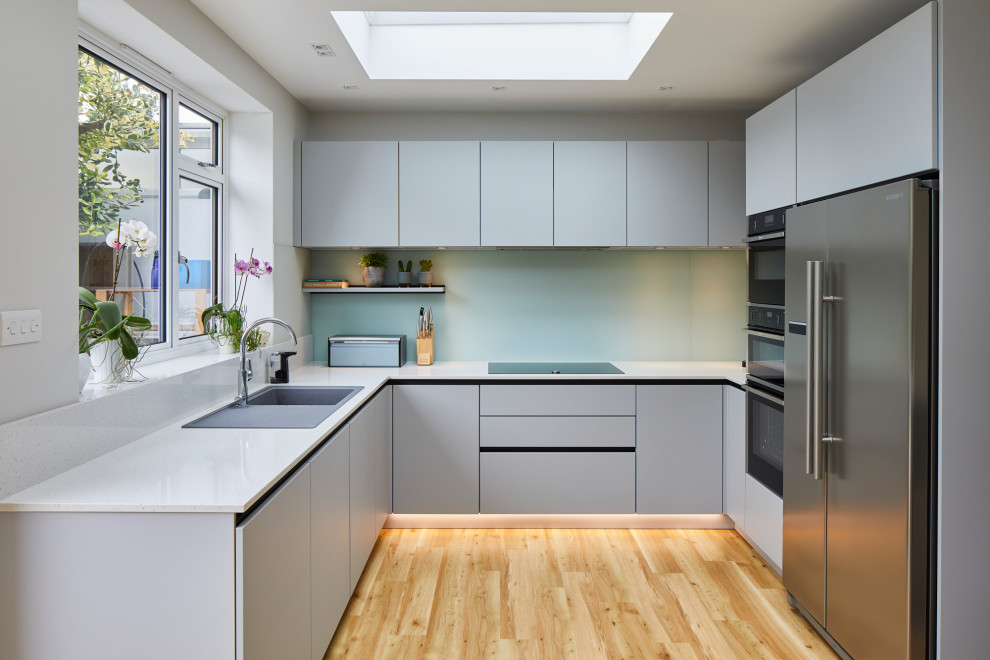Inspiration for a mid-sized contemporary u-shaped eat-in kitchen in London with flat-panel cabinets, grey cabinets, quartzite benchtops, green splashback, glass sheet splashback, stainless steel appliances, a peninsula and white benchtop.