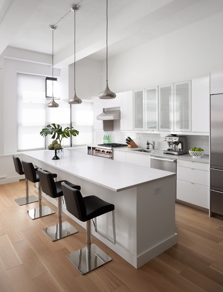 This is an example of a mid-sized modern single-wall open plan kitchen in New York with an undermount sink, flat-panel cabinets, white cabinets, quartz benchtops, white splashback, ceramic splashback, stainless steel appliances, light hardwood floors, with island and beige floor.