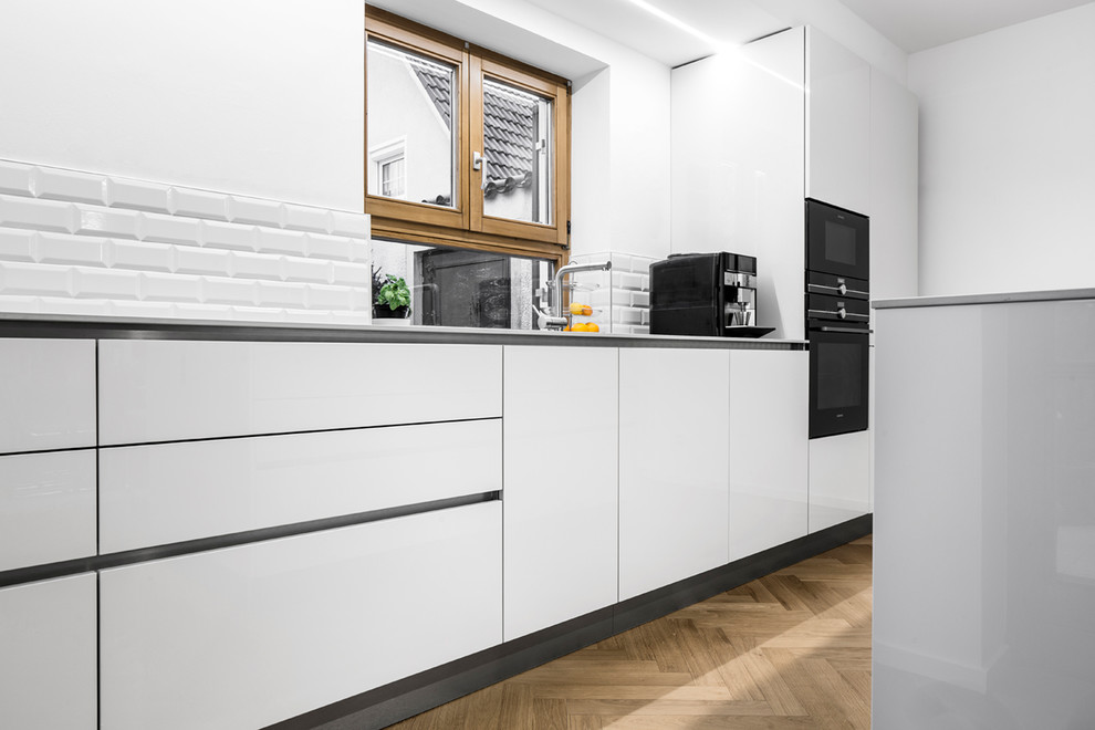 Zweizeilige, Große Moderne Wohnküche mit Waschbecken, flächenbündigen Schrankfronten, grauen Schränken, Mineralwerkstoff-Arbeitsplatte, Küchenrückwand in Weiß, Rückwand aus Metrofliesen, schwarzen Elektrogeräten, hellem Holzboden, Kücheninsel, braunem Boden und grauer Arbeitsplatte in München