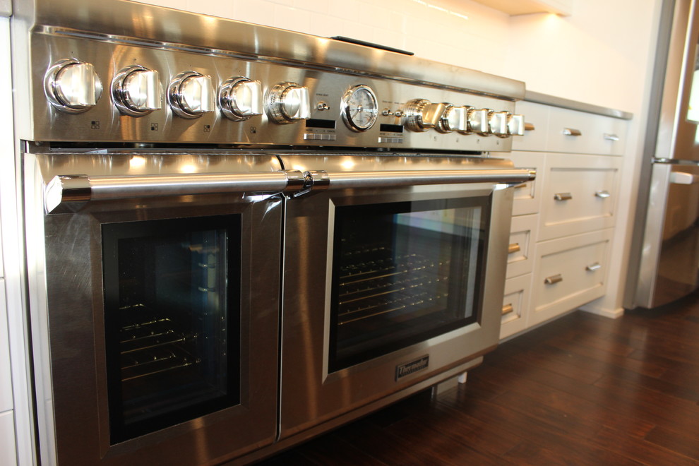 Example of a large transitional l-shaped dark wood floor enclosed kitchen design with a farmhouse sink, shaker cabinets, white cabinets, quartz countertops, white backsplash, subway tile backsplash, stainless steel appliances and an island